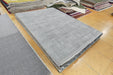 A rectangular light gray rug sample or finished rug shown on a wood floor, with stacks of folded rugs in the background.
