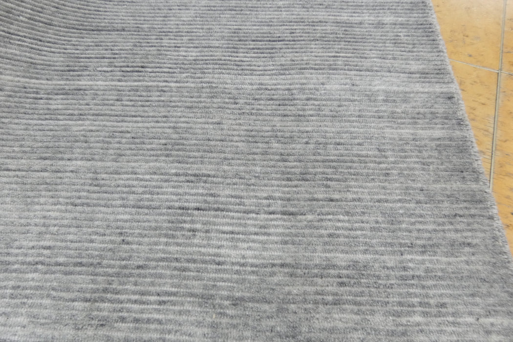 A close-up of a gray textured rug with horizontal ribbed stripes, shown with a small portion of a light wood floor visible on the right edge.