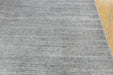 A close-up of a gray textured rug with horizontal ribbed stripes, shown with a small portion of a light wood floor visible on the right edge.