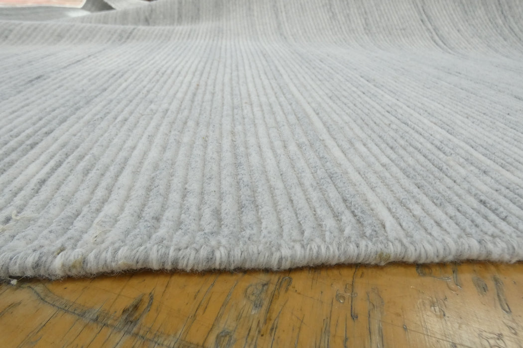 A close-up of a light gray ribbed rug or carpet laid over a wooden floor, showing the texture and edge of the textile.