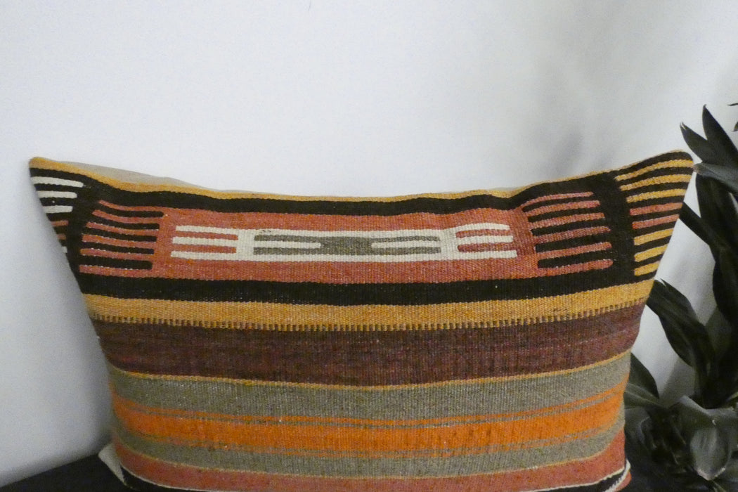 A rectangular kilim throw pillow with a Southwestern-style woven pattern in orange, brown, beige, and black, shown against a white wall with a plant leaf visible on the right.