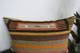 A rectangular kilim throw pillow with a Southwestern-style woven pattern in orange, brown, beige, and black, shown against a white wall with a plant leaf visible on the right.