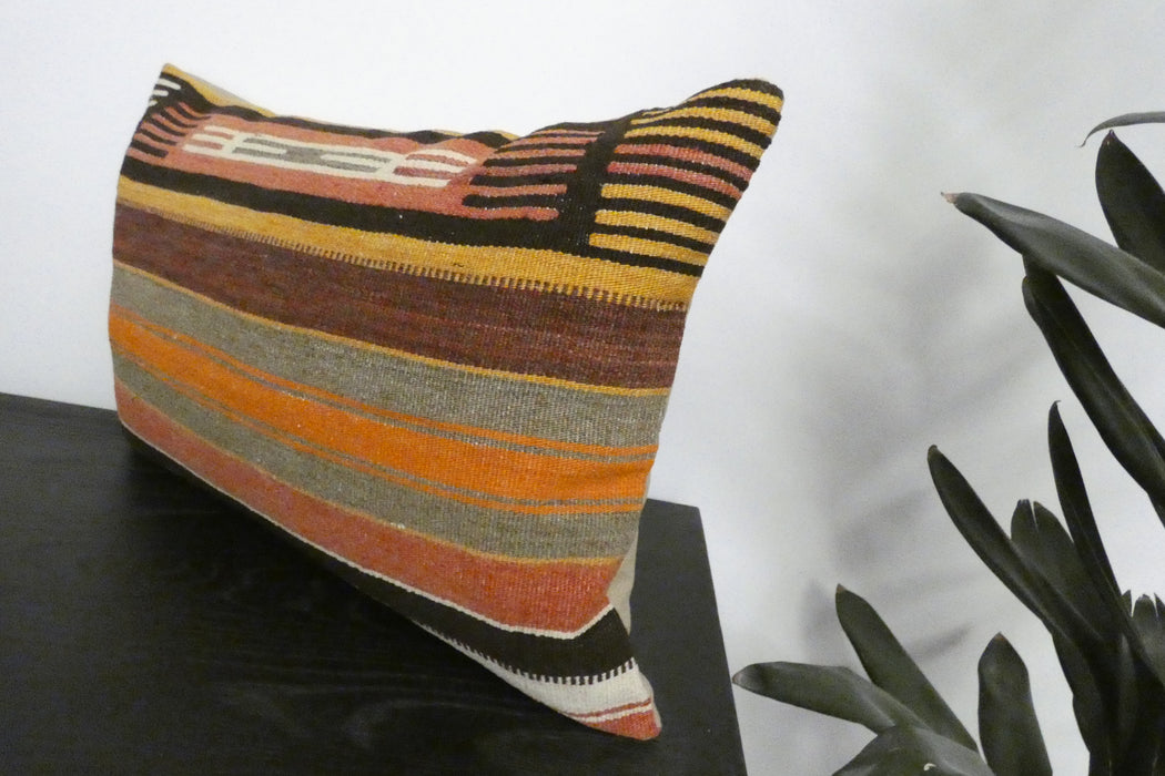 A square throw pillow with a rustic woven kilim pattern featuring horizontal stripes of orange, brown, beige, black, and white, resting on a dark wooden surface next to a green plant against a white wall.