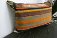 A square woven throw pillow with horizontal stripes in orange, brown, beige, black, and white, resting on a dark wooden surface against a white wall.