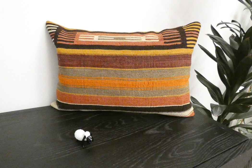 A rectangular woven throw pillow with horizontal stripes in orange, brown, beige, and black, resting on a dark wooden table next to a green plant.