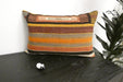 A rectangular woven throw pillow with horizontal stripes in orange, brown, beige, and black, resting on a dark wooden table next to a green plant.