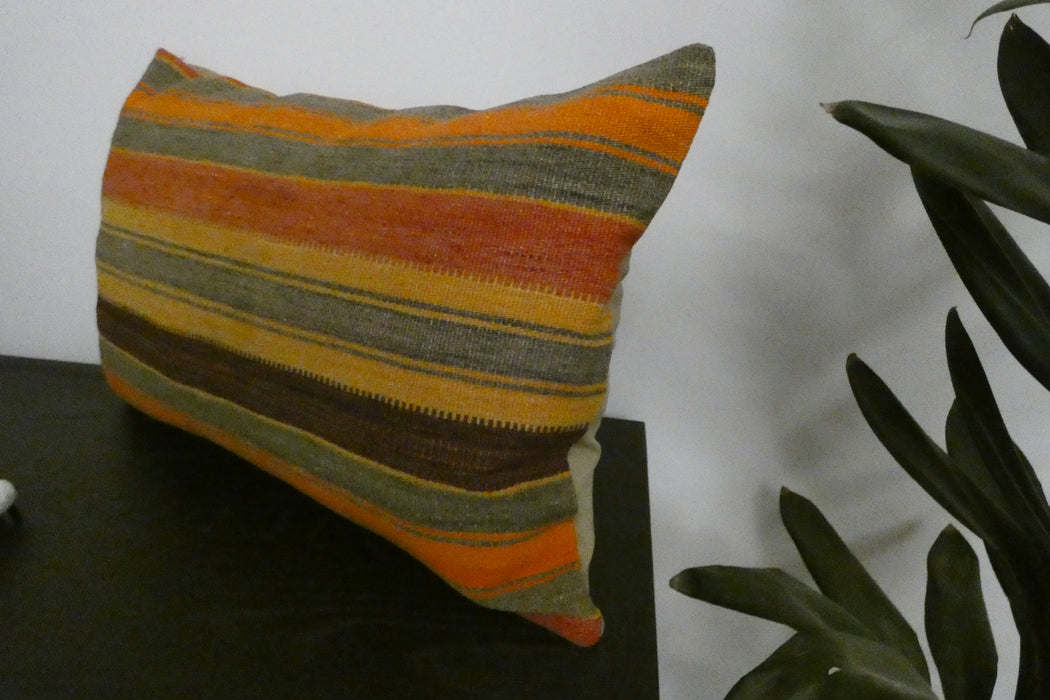 A rectangular throw pillow with a rustic striped pattern in orange, yellow, brown, and gray, resting on a dark table next to a green plant.