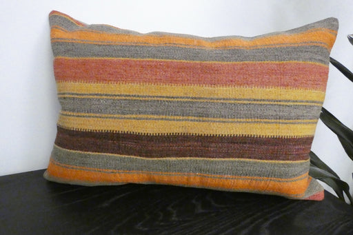 A rectangular woven throw pillow with horizontal stripes in orange, yellow, brown, and gray, resting on a dark wooden surface against a white wall.