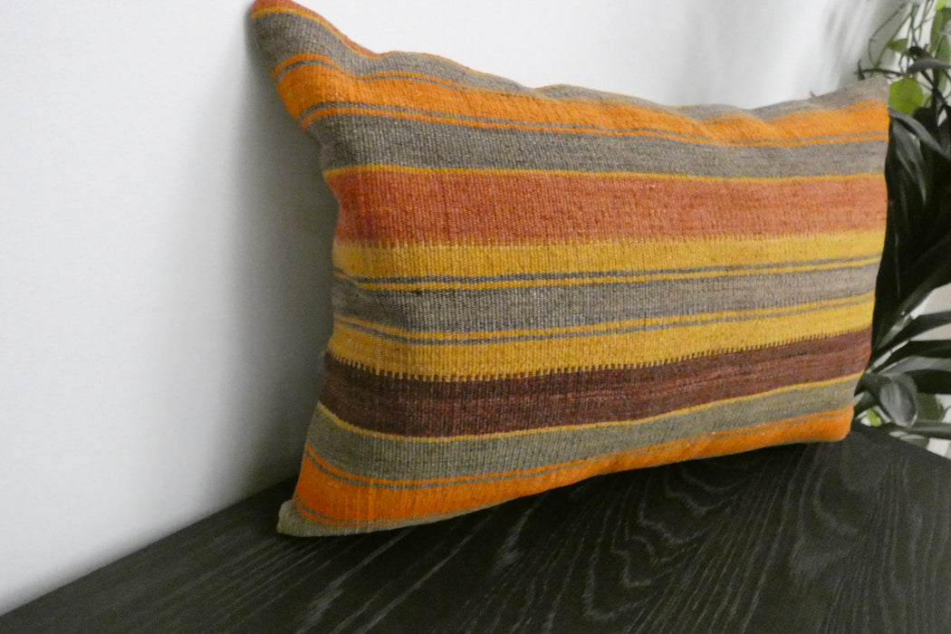 A rectangular woven throw pillow with horizontal stripes in orange, yellow, brown, and gray, resting on a dark wooden surface against a white wall.