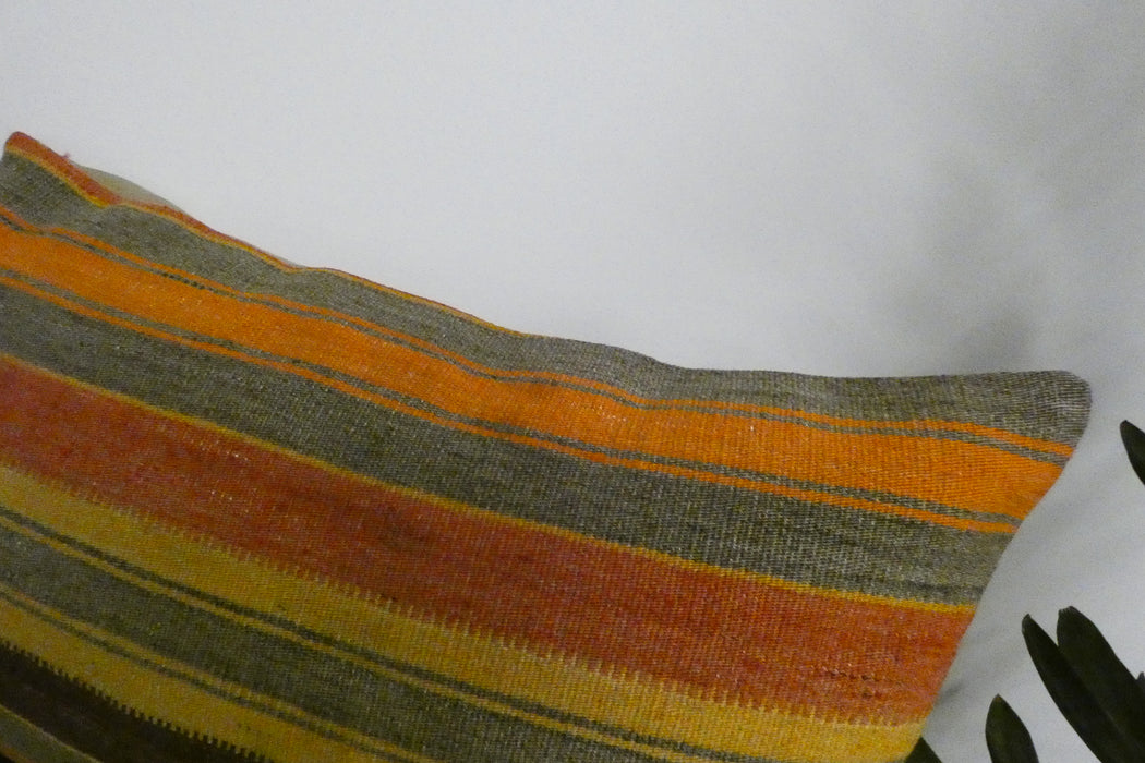 A close-up of a woven throw pillow featuring horizontal stripes in orange, yellow, brown, and gray, set against a plain white background with a plant leaf visible in the corner.