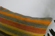 A close-up of a woven throw pillow featuring horizontal stripes in orange, yellow, brown, and gray, set against a plain white background with a plant leaf visible in the corner.