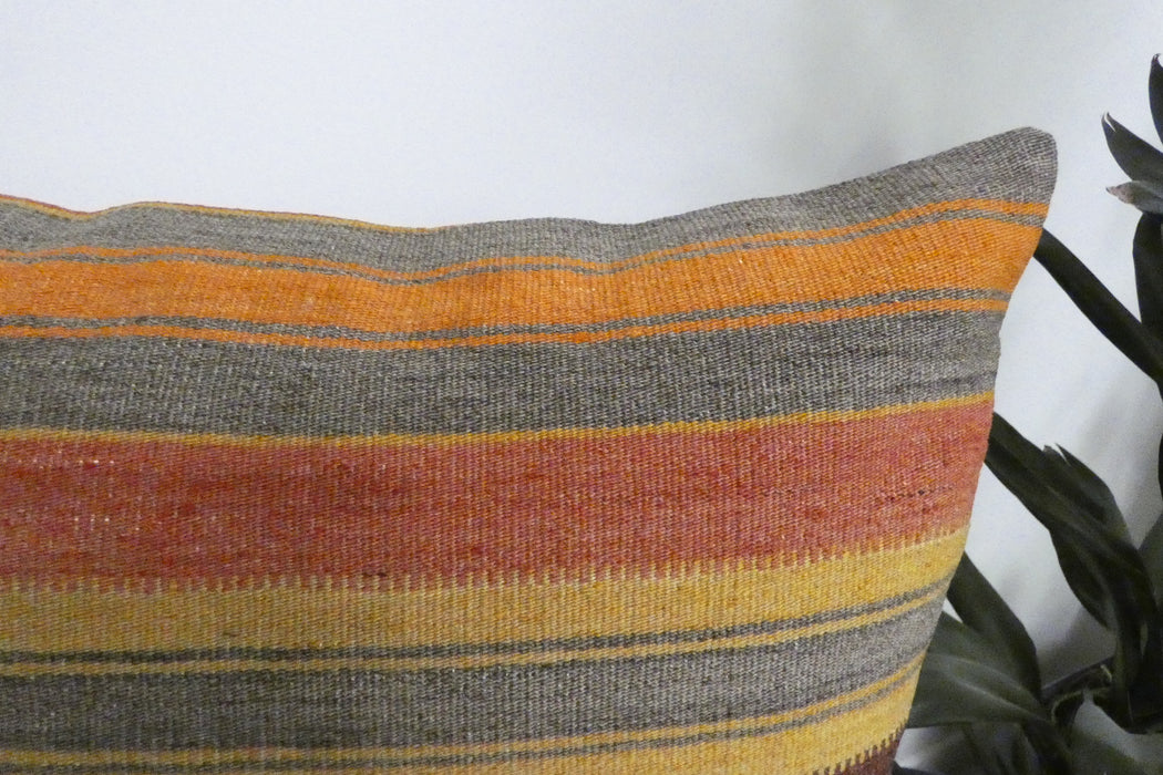 A close-up of a woven throw pillow with horizontal stripes in gray, orange, rust red, and yellow, shown next to a green plant against a white wall.