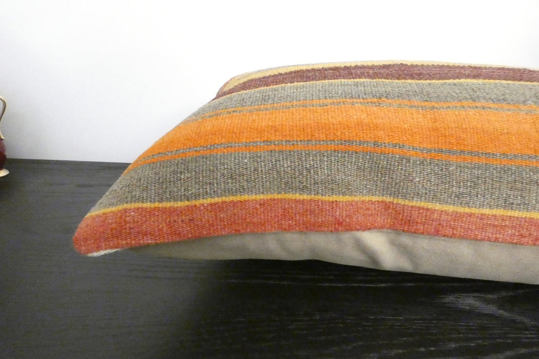 A close-up of a striped throw pillow with a textured woven fabric in orange, brown, gray, and rust tones, resting on a dark wooden surface.