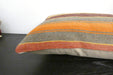 A close-up of a striped throw pillow with a textured woven fabric in orange, brown, gray, and rust tones, resting on a dark wooden surface.