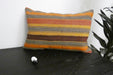 A rectangular woven throw pillow with horizontal stripes in orange, yellow, brown, and gray, resting on a dark wooden table next to a small white sheep figurine and a green plant.