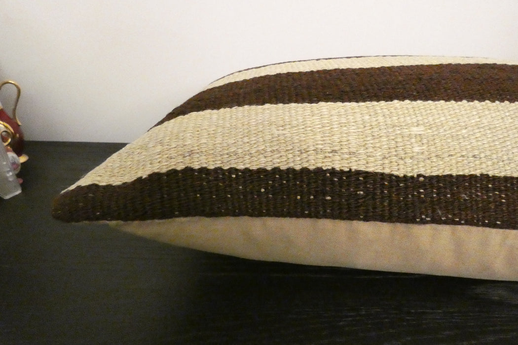 A rectangular woven throw pillow with horizontal stripes in dark brown and cream, resting on a dark wooden surface against a plain white wall.