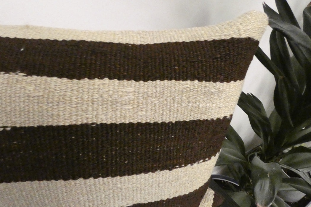 A close-up of a woven throw pillow with horizontal stripes in dark brown and cream, positioned next to a green houseplant.
