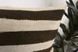A close-up of a woven throw pillow with horizontal stripes in dark brown and cream, positioned next to a green houseplant.